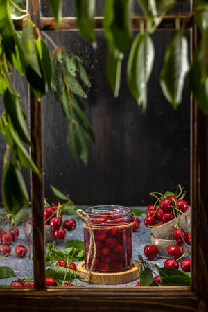 Jar of homemade cherry jam is sitting on a window sill, surrounded by fresh cherriesの写真素材