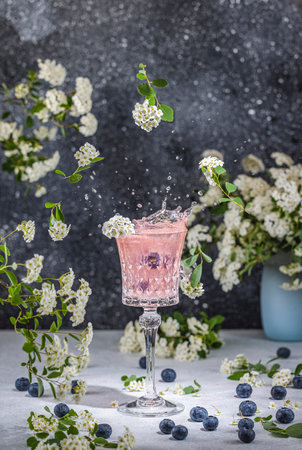Pink cocktail splashing in a crystal glass, garnished with white flowers and blueberries on a tableの写真素材