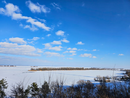 Scenic winter landscape featuring a frozen river stretching to the horizon under a vibrant blue sky dotted with fluffy white cloudsの写真素材