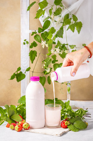 Hand pouring strawberry yogurt from bottle into glass, with fresh strawberries, mint, and decorative plants, promoting healthy eating and refreshmentの写真素材