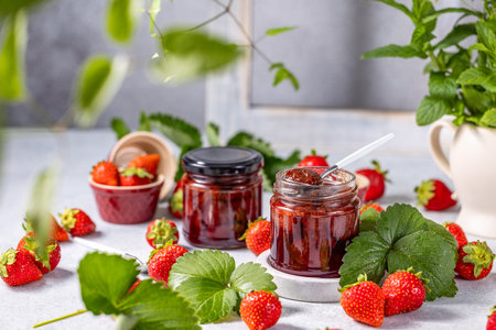 Two jars of strawberry jam, one with a spoon inside, surrounded by fresh strawberries, leaves, and a mint plantの写真素材