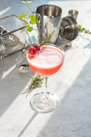 Pink cocktail decorated with raspberries and rosemary sprig, stands on bright table with cocktail shaker and jigger in backgroundの写真素材