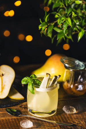 Pear cocktail with basil and fruit slices is served on a wooden table with ice cubes, a spoon, and a jar of pear jam in the background with bokeh effectの写真素材