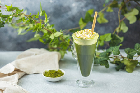 Green tea powder and ivy branches near a tall glass of iced matcha latte with whipped cream and a...