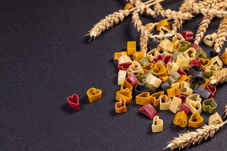 Small colorful heart shaped pasta is arranged on a black background with wheat, creating a romantic and culinary sceneの写真素材