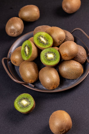 Ripe kiwi fruits are arranged on a metal tray and scattered on a dark surface, showcasing their fuzzy brown skin and juicy green fleshの写真素材
