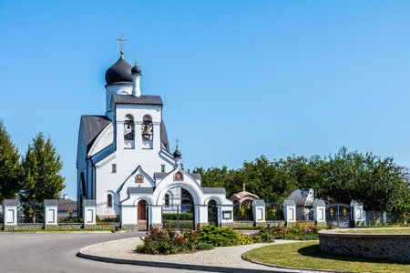 White orthodox church with black domes and golden crosses, standing tall against a clear blue sky, surrounded by green trees and a paved areaの写真素材