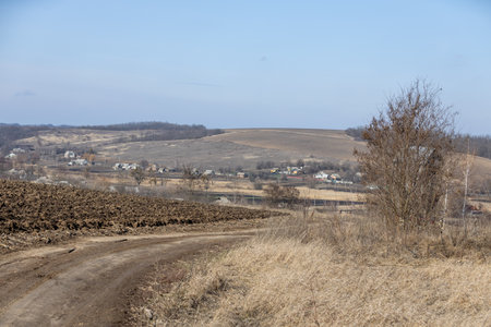 Plowed field and dirt road leading to a small village in the countryside, with dry grass and bare treesの写真素材