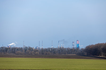 Smoke rising from an industrial plant behind a green field and forest, highlighting environmental impactの写真素材