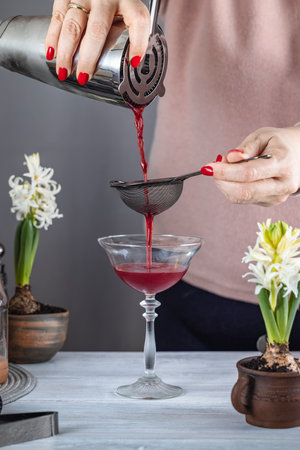 Bartender filtering red cocktail from shaker through strainer into a coupe glass, white hyacinth flowers on a gray backgroundの写真素材
