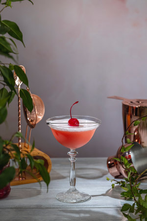 Refreshing pink cocktail with egg white foam, garnished with a maraschino cherry, served in a coupe glass on a wooden table with bar tools and plantsの写真素材