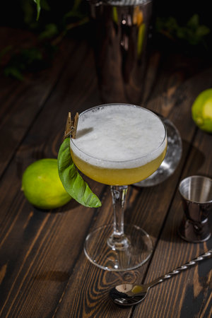 Vibrant yellow cocktail topped with white foam sits in a coupe glass on a dark wooden table, garnished with lime and leafの写真素材