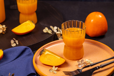 Glass of vibrant persimmon juice accompanied by fresh persimmon slices on a stylish plate, creating a sophisticated table settingの写真素材