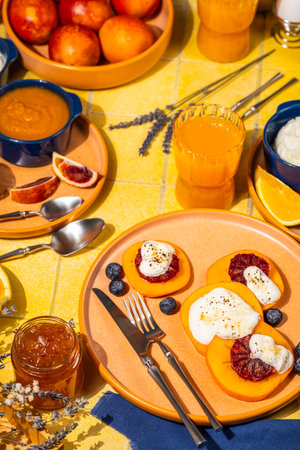 Vibrant breakfast featuring persimmon slices topped with ricotta, blood orange, and blueberries on a colorful table with juice and jamの写真素材