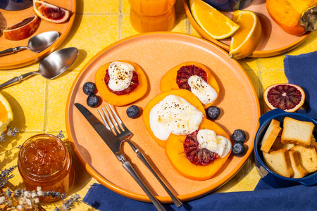 Delicious persimmon dessert with ricotta cheese, blood orange slices and blueberries, served on a vibrant table with orange juice, jam, and melba toastの写真素材