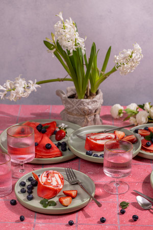 Red velvet pancakes and cream cheese dessert rolls are served on a pink tiled table with pink drinks, strawberries, blueberries, and hyacinth flowersの写真素材