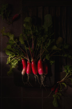 Vibrant red radishes with lush green leaves resting on a dark cutting board create a striking contrast against a rustic backdrop, highlighting fresh, farm to table ingredientsの写真素材