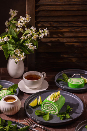 Green lime matcha roll cakes with cream and fresh lime slices on plates are ready for serving with hot black tea and delicate white jasmine flowers on a dark backgroundの写真素材