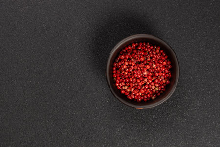 Pink peppercorns, a delicate spice and cooking ingredient, filling a small dark bowl on a black background, offering a natural and elegant seasoning optionの写真素材