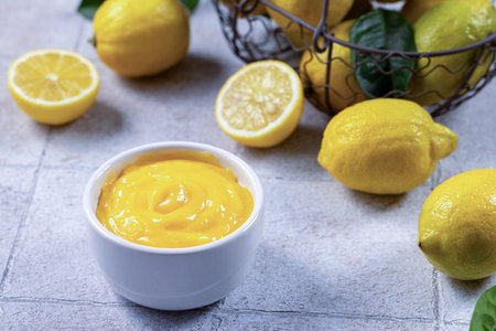 Creamy yellow lemon curd in a white bowl with fresh whole and halved lemons, showing bright zest and smooth texture for baking, breakfast or dessert preparation, overhead studio shotの写真素材