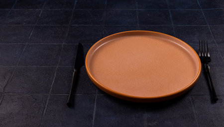 Empty ceramic dinner plate with a black knife and fork resting on a dark, textured tabletop,...