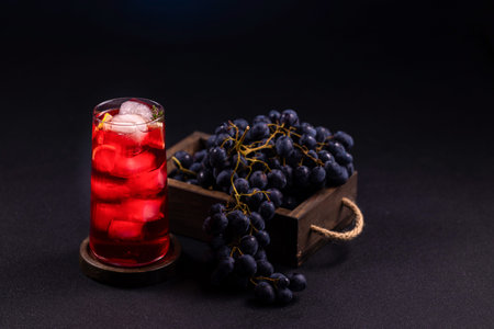 Tall glass containing a vibrant red cold drink with ice cubes and lemon slices, presented alongside dark purple grapes overflowing from a rustic wooden box on a dark backgroundの写真素材