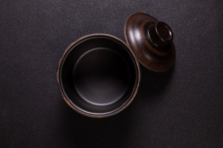 Rustic ceramic bowl with matching lid separated on textured dark surface, overhead flat lay...