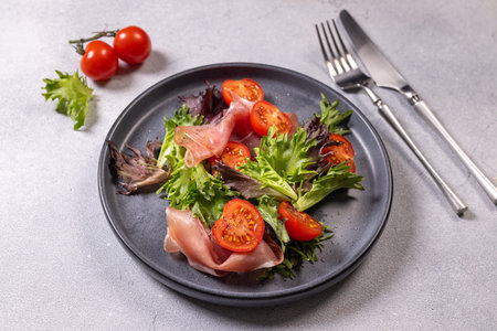 Fresh green salad with vibrant cherry tomatoes and delicate prosciutto slices arranged on a dark plate, presenting a healthy and delicious appetizer or light mealの写真素材
