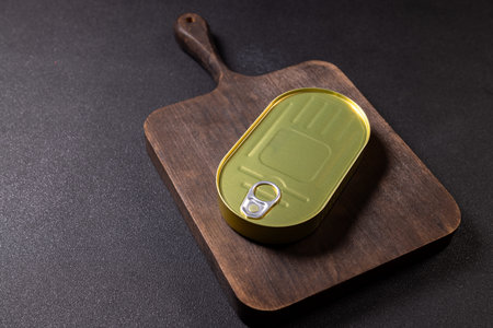 Oval, golden green tin of preserved food with a pull ring tab resting on a dark wooden cutting board, presenting a simple still life composition with a black backgroundの写真素材