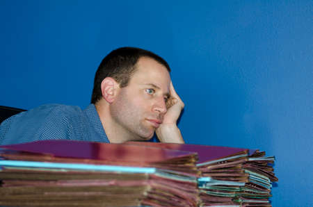 Man worn out at work looking straight ahead sitting in front of a large pile of files with his head leaning on his hand.の写真素材