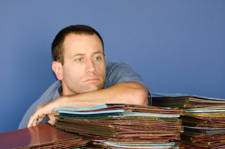 Man stressed out at work leaning on a large pile of files while staring straight ahead.