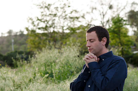 Man praying alone outside in a grassy field with hands folded and eyes closed.の写真素材