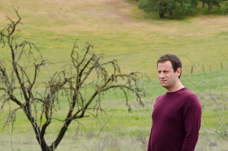 Man in deep thought in an open grassy field with a bare tree in the background.の写真素材