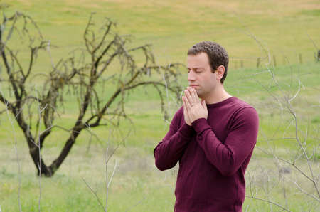 Man praying outside in an empty field with a bare tree in the background.の写真素材