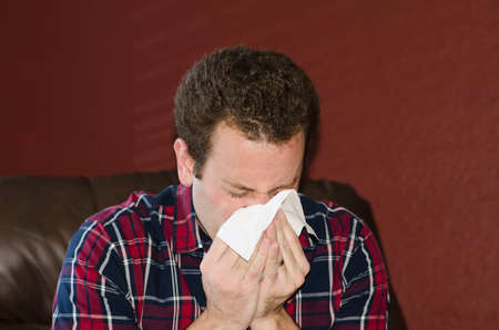 Sick man at home blowing his nose into a tissue sitting in a leather chair.の写真素材