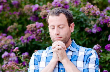 Close up of man praying alone outside with eyes closed and hands clasped in front of purple flowers.の写真素材