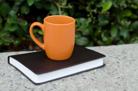 Orange mug and black leather bound book outside on a cement ledge with bushes in the background.の写真素材