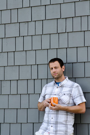 Man leaning against a gray wall holding an orange coffee mug outside.の写真素材