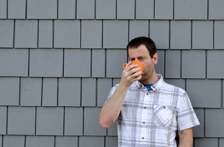Man drinking from an orange coffee mug against a gray background.の写真素材