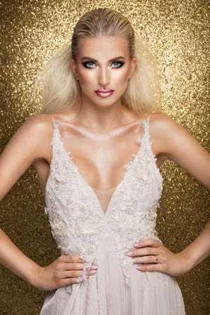 Sensual beautiful blonde woman posing in white dress over gold background. Girl with long curly hair.の写真素材