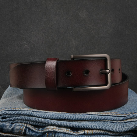 A stylish dark red leather belt with a silver buckle, placed on a pair of folded blue jeans against...