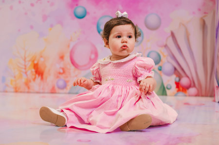 Cute baby girl in pink dress sitting on the floor and looking at cameraの写真素材