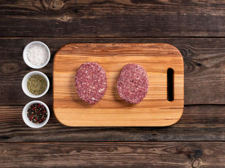 Raw organic meat cutlets for hamburgers on wooden cutting board. Plates with seasonings. Top view.の写真素材