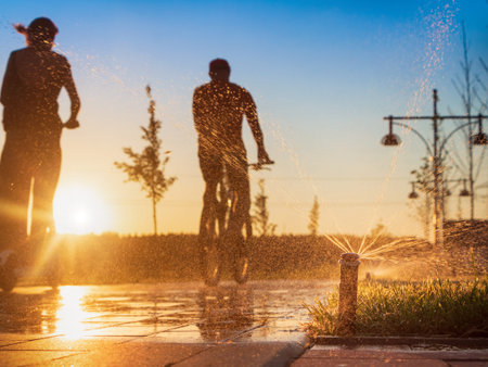 Automatic sprinkler system watering the lawn on a background of man on bike and girl on scooter riding in public park at sunset.の写真素材