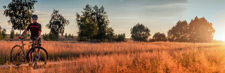 Beautiful panorama of a guy with a gravel bike standing on a path in a field at sunset. Beautiful...