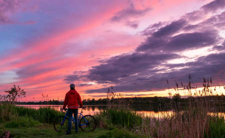 Cyclist on the coast of the river at sunset in summer. Landscape with man on gravel bike and colorful sky with clouds. Sport and travel concept.の写真素材