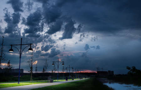 The path of the city park near the river with lanterns against the sky with rain clouds.Beautiful city landscape at sunset.の写真素材