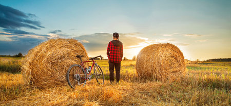 A dramatic landscape of a cyclist standing between two haystacks in a field at sunset. Active...