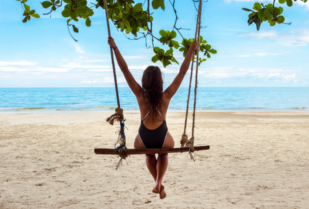 Young girl sits on a swing on a sandy beach by the ocean and enjoys the view.の写真素材