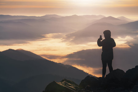 Young girl on the mountain peak photographs a beautiful mountain landscape at sunrise on the phone.